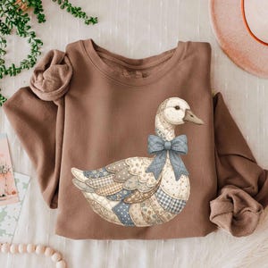 Pode incluir: Um moletom marrom com um design de ganso de retalhos e um laço azul. O ganso tem uma cabeça branca e um bico cinza. O design de retalhos incorpora padrões florais, de bolinhas e xadrez em tons de azul, marrom e bege.