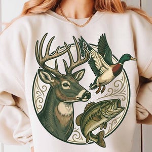 Puede incluir: Sudadera color crema con un diseño gráfico circular que representa un ciervo, un pato y un pez. El ciervo tiene grandes cuernos, el pato está en vuelo y el pez tiene la boca abierta. El diseño utiliza tonos verdes, marrones y beige.
