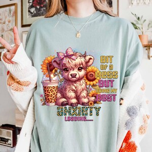 May include: A light blue t-shirt featuring a cartoon Highland cow with a pink bow, sunflowers, and a leopard-print drink. Text on the shirt reads "Bit of a mess but doing my best" and "Anxiety Loading".