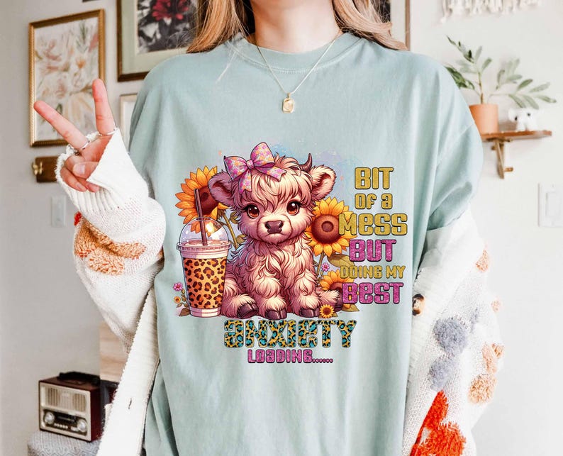 May include: A light blue t-shirt featuring a cartoon Highland cow with a pink bow, sunflowers, and a leopard-print drink. Text on the shirt reads "Bit of a mess but doing my best" and "Anxiety Loading".