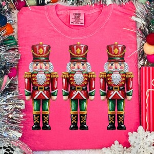 May include: A bright pink Comfort Colors t-shirt featuring three detailed nutcracker soldier illustrations. The nutcrackers are in red, green, and gold, with white beards and black boots. The shirt has a crew neck and short sleeves.