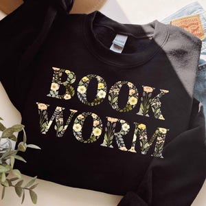 Peut inclure: Sweat-shirt noir avec l'inscription "BOOK WORM" dans un motif floral. Les lettres sont remplies de fleurs jaunes, blanches et vertes. Le sweat-shirt est présenté avec un jean en denim.