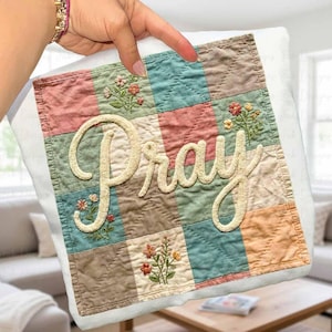 May include: A square, quilted pillow with a patchwork design in pastel colors. The word "Pray" is embroidered in a cursive font across the center. Small floral embroidery accents the patchwork.