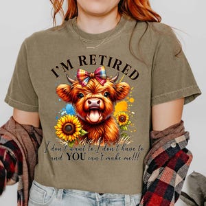 May include: Olive green t-shirt featuring a watercolor illustration of a Highland cow with a colorful bow, sunflowers, and the text "I'M RETIRED". The shirt also includes the text "I don't want to, I don't have to and YOU can't make me!!!".