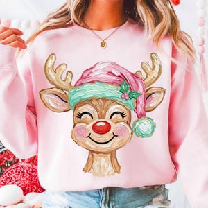 May include: Pink sweatshirt featuring a watercolor illustration of a reindeer wearing a pink and green Santa hat. The reindeer has a red nose, rosy cheeks, and a smiling expression. The antlers are gold.