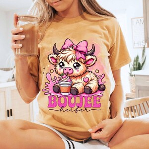 May include: Mustard yellow t-shirt featuring a cartoon cow with a pink bow, drinking a beverage. The text "BOUJEE heifer" is printed in pink. The cow has large eyes and is surrounded by pink splashes and stars.