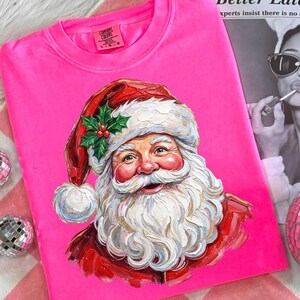 May include: A bright pink Comfort Colors sweatshirt featuring a colorful illustration of Santa Claus. Santa is wearing a red hat with holly and berries, and a white beard. The sweatshirt is a festive holiday item.