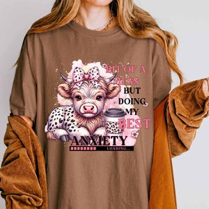 May include: Brown t-shirt with a cartoon cow and a coffee cup. The text on the shirt reads "Bit of a mess but doing my best anxiety loading..."