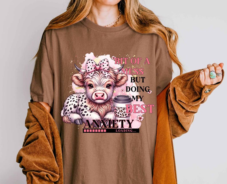 May include: Brown t-shirt with a cartoon cow and a coffee cup. The text on the shirt reads "Bit of a mess but doing my best anxiety loading..."