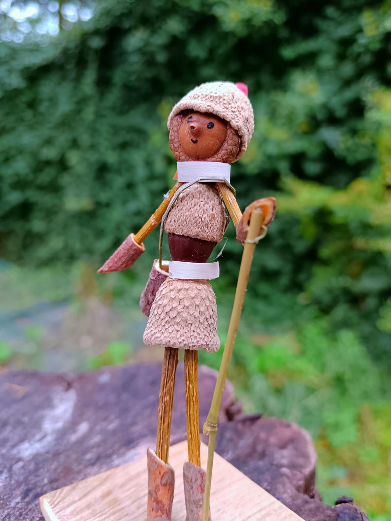 May include: A whimsical figurine made from natural materials, featuring a small person with a brown acorn head, wearing a brown knitted hat with a red pom-pom, a brown skirt, and a white scarf. The figurine is holding a stick and standing on a wooden base.