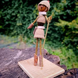 May include: A whimsical figurine made from natural materials, such as acorns and twigs, is dressed in a brown and white outfit with a red and white hat. The figurine is holding a stick and standing on a wooden base.