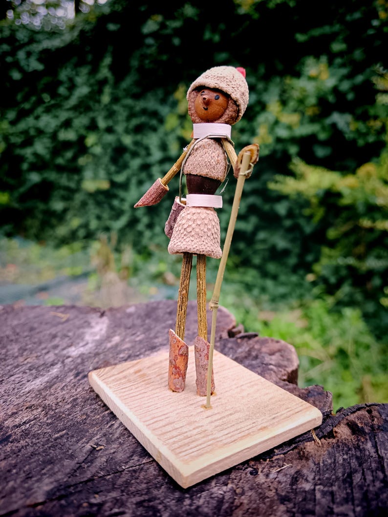 May include: A whimsical figurine made from natural materials, such as acorns and twigs, is dressed in a brown and white outfit with a red and white hat. The figurine is holding a stick and standing on a wooden base.