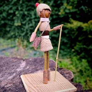 May include: A whimsical figurine made from natural materials, featuring a stick figure with a brown acorn head, a red hat, and a brown acorn skirt. The figure is holding a stick and is standing on a wooden base.