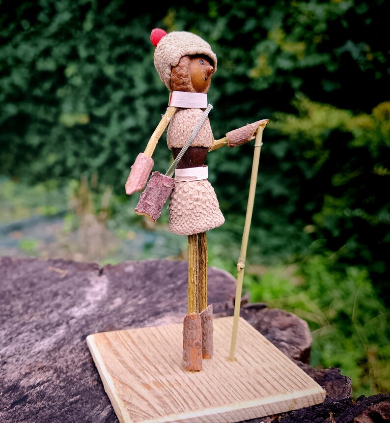 May include: A whimsical figurine made from natural materials, featuring a stick figure with a brown acorn head, a red hat, and a brown acorn skirt. The figure is holding a stick and is standing on a wooden base.