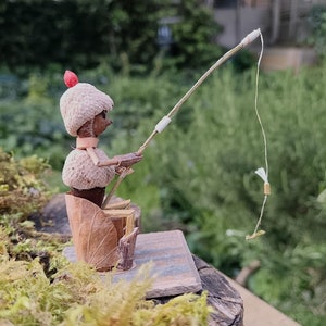 Puede incluir: Una caprichosa figura en miniatura de un pescador hecha con materiales naturales. La figura, hecha de bellotas y hojas, está sentada y sostiene una caña de pescar con un hilo delicado. La escena está ambientada al aire libre con un fondo verde borroso.
