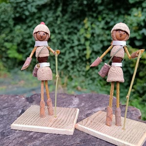 May include: Two small, whimsical figures made from natural materials like acorns and twigs. They are dressed in brown and white and wear hats. Each figure holds a stick and stands on a small wooden base.