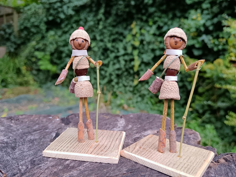 May include: Two small, whimsical figures made from natural materials like acorns and twigs. They are dressed in brown and white and wear hats. Each figure holds a stick and stands on a small wooden base.