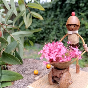 May include: A small, whimsical figurine made from natural materials, depicting a person with a red hat and a white bow tie, pushing a wheelbarrow filled with pink flowers. The figurine is sitting on a wooden base.