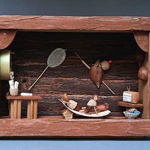 Pode incluir: Um diorama em caixa de madeira com figuras em miniatura feitas de bolotas e materiais naturais. A cena inclui uma pequena mesa, uma espreguiçadeira, um televisor e uma rede na parede, tudo dentro de uma moldura castanha rústica.
