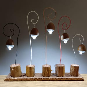 May include: Six small, brown, acorn-shaped lights with white LED bulbs are suspended from wire stems on a wooden base. The stems are wrapped in black, white, and red wire.