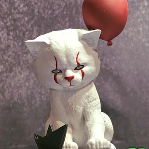 May include: White cat figurine with red clown makeup and a red balloon. The cat is sitting on a black base with a paper boat.