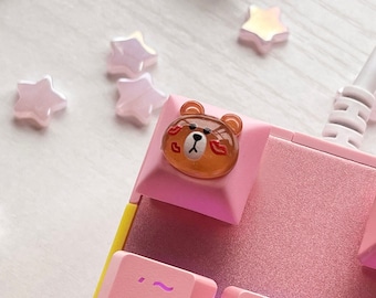 May include: A pink keyboard with a brown bear keycap. The bear has a red heart and a red lipstick mark on its face. The keyboard has pink keys with white lettering. The keys are labeled with numbers, letters, and symbols. The keyboard is on a white surface.