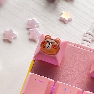 May include: A pink keyboard with a brown bear keycap. The bear has a red heart and a red lipstick mark on its face. The keyboard has pink keys with white lettering. The keys are labeled with numbers, letters, and symbols. The keyboard is on a white surface.