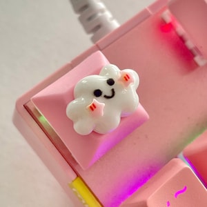 May include: A pink mechanical keyboard with a white cloud keycap featuring a smiling face and pink stars. The keyboard has a rainbow backlight.