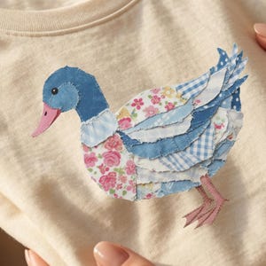 May include: Cream-colored t-shirt featuring a patchwork duck design. The duck has a blue head, pink beak, and wings made of floral and checkered fabric. The shirt is folded and held in someone's hands.