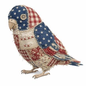 Op de afbeelding: Een patriottische stoffen vogelbeeldje met een patchwork-ontwerp. De vogel heeft rode, witte en blauwe stoffen met ster- en bloempatronen. Een knoop dient als oog en de snavel is beige. De vogel staat op twee gedetailleerde voeten.