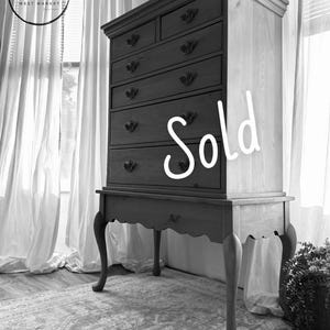 May include: A tall wooden chest of drawers featuring six drawers with dark ornate metal pulls, resting on a base with four curved cabriole legs and a scalloped apron. The word Sold is written in white script across the front of the drawers.