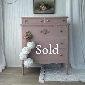 May include: A dusty rose painted wooden dresser featuring four drawers with round brass knobs, a decorative carved molding detail on the second drawer, and a hanging cluster of white fabric flowers tied with a tan ribbon on the left side.