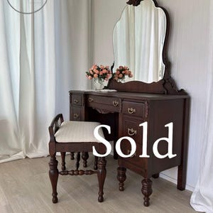 May include: A dark wood vanity table featuring a large arched mirror, multiple drawers with brass hardware, and a matching stool with a light-colored upholstered seat. The word Sold is printed in white across the center of the furniture.