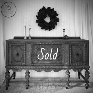 May include: A dark wood antique sideboard with ornate carvings, featuring the word "Sold" in script. Two silver candlesticks and a black wreath adorn the top. The West Market logo is in the upper left corner.