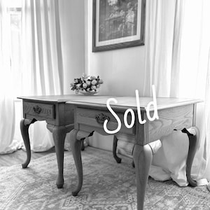 May include: A pair of wooden side tables featuring a single drawer with ornate dark metal hardware and curved cabriole legs. The tables have a visible wood grain texture and are marked with the word Sold in white script across the front.