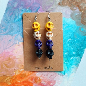 May include: Dangle earrings with a stacked skull design. Each earring has a yellow, white, purple, and black skull. The earrings are displayed on a brown card. The text "aus_asche" is visible on the card.