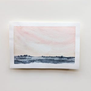 May include: A rectangular watercolor painting on textured white paper featuring a landscape with a dark blue, horizontal band representing land and trees against a soft, pale pink and peach gradient sky that fills the upper portion.