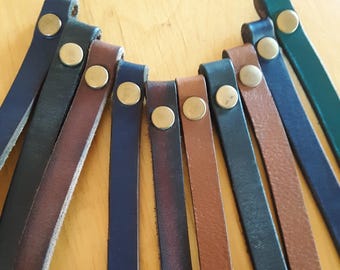 Puede incluir: Primer plano de correas de cuero en varios colores, incluyendo azul marino, marrón y verde. Cada correa está asegurada con un broche de color latón. Las correas están dispuestas en un patrón semicircular, sugiriendo un collar u otro accesorio.