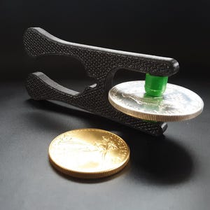 May include: A black 3D-printed coin holder with a textured surface, holding a silver coin. A gold coin rests below. The holder has a green cylindrical piece. The background is black.