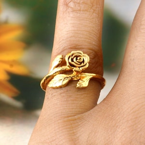 Könnte beinhalten: Ein goldfarbener Ring mit einem Rosendesign. Der Ring hat eine detaillierte Rosenblüte und Blätter, mit einem Band, das sich um den Finger legt. Der Ring wird an einem Finger gezeigt.