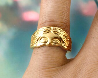 May include: A gold-colored ring featuring a sculpted face design. The ring's face has detailed features, including eyes and flowing hair. The ring is worn on a finger, showcasing its intricate craftsmanship and design.