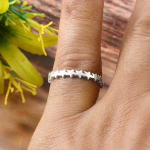 May include: A silver ring with a repeating star pattern. The ring is on a finger, with a blurred background of yellow flowers and wood. The ring's design features a series of small, connected star shapes.