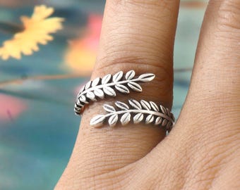 May include: A silver-colored ring wraps around a finger, featuring a detailed leaf design. The ring's leaves are arranged in a spiral pattern, creating an elegant and natural aesthetic. The ring is a piece of jewelry.