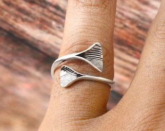 Puede incluir: Un anillo plateado con un diseño único. El anillo presenta dos elementos en forma de abanico con líneas detalladas, creando una apariencia texturizada. El anillo se envuelve alrededor del dedo, ofreciendo un ajuste ajustable. El anillo se muestra en el dedo de una persona.
