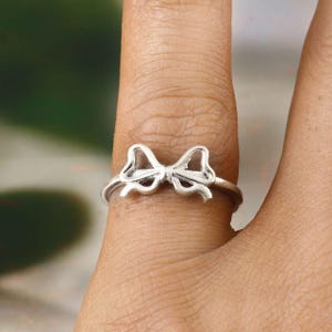 May include: A silver ring with a bow design. The ring is a delicate piece of jewellery, with the bow's details clearly visible. The ring is worn on a finger, showcasing its size and style. This ring is a charming accessory.
