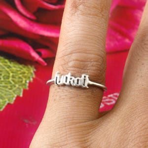 Puede incluir: Anillo de plata con la palabra "fuckoff" en cursiva. El anillo está en un dedo, con un fondo borroso de una flor roja.