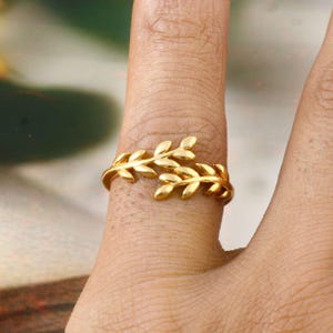 May include: A gold-colored ring featuring a leaf design. The ring wraps around the finger with two detailed leaf branches. The ring is made of a shiny, metallic material.