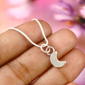 May include: A silver necklace featuring a crescent moon pendant. The pendant is attached to a delicate silver chain. The necklace is displayed against a soft, blurred background, highlighting the jewelry's design.