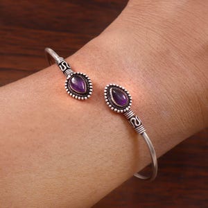 Può includere: Un bracciale rigido in argento con due pietre preziose di ametista viola a forma di goccia alle estremità aperte. La fascia in metallo presenta intricati dettagli ossidati e un bordo perlato attorno a ogni pietra per un aspetto strutturato.