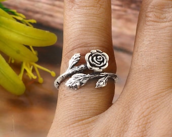 Può includere: Un anello color argento con un design a rosa. L'anello presenta un fiore di rosa e foglie dettagliate, avvolti attorno al dito. L'anello è indossato su un dito, con uno sfondo sfocato.